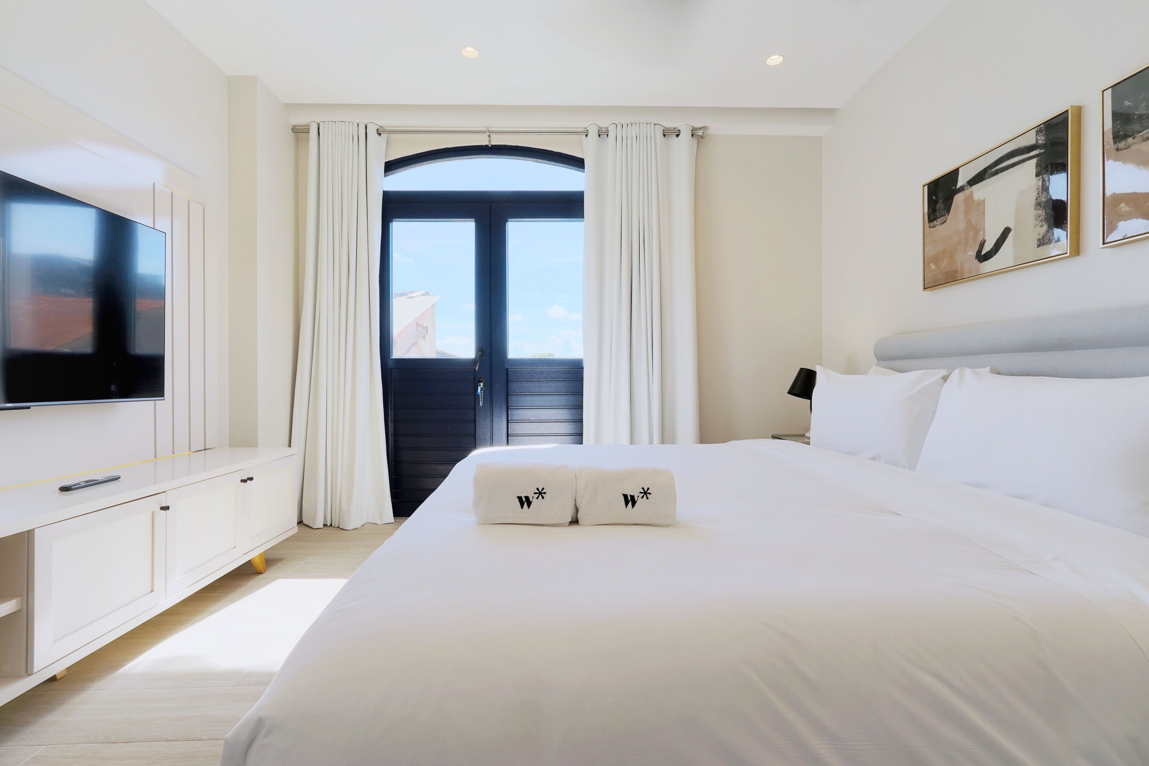 A modern hotel room with a large bed, a flat-screen TV, a desk, and a balcony with curtains.