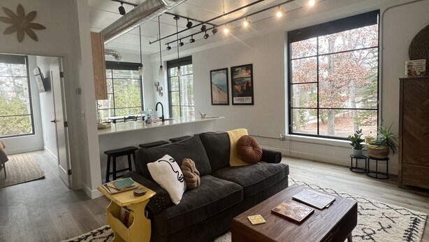 A modern living room with a dark sofa, a wooden coffee table, and a patterned rug. There is a large window with a view of trees and a kitchen area in the background. - Cozy, Eclectic 1 BR Condo w/Rooftop Hot Tubs, Dog Friendly! (Traverse City)
