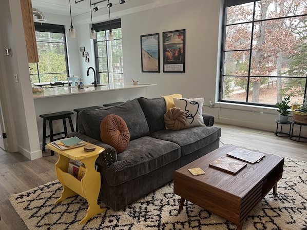 A modern living room with a dark grey sofa, a wooden coffee table, and a yellow side table. There are two framed pictures on the wall, a window with a view of trees, and a kitchen area in the background.