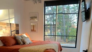 A bedroom with a large window, a bed with orange bedding, a wooden floor, and a ceiling light fixture. - Cozy, Eclectic 1 BR Condo w/Rooftop Hot Tubs, Dog Friendly! (Traverse City)