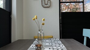 A room with a large window, a table with a patterned tablecloth, a vase with dried flowers, and two wooden letters 'TC' on the wall. - Cozy, Eclectic 1 BR Condo w/Rooftop Hot Tubs, Dog Friendly! (Traverse City)