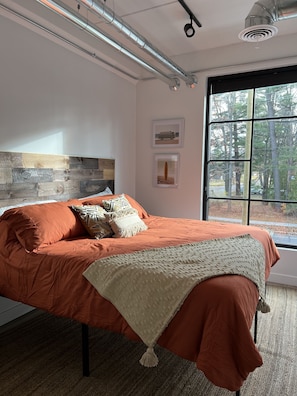 A bedroom with a bed, a headboard, a window with a view of trees, and a ceiling with ductwork.