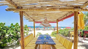 Un área de comedor al aire libre con una pérgola de madera, una mesa con sillas y vista a la playa y al océano.