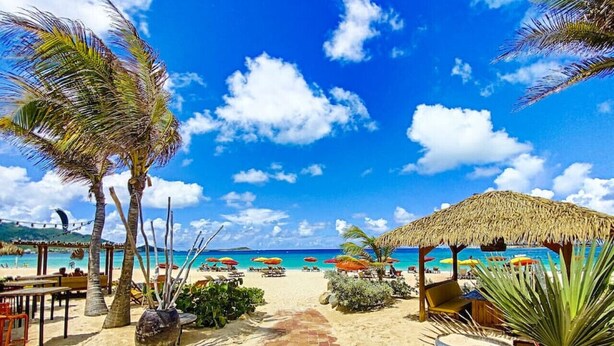 Un sendero bordeando la playa que conduce a una zona con sombrillas, palmeras y una estructura de techo de paja.
