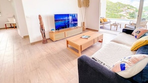 Ein modernes Wohnzimmer mit großem Fernseher, Holzmöbeln und Blick auf eine Hügellandschaft.