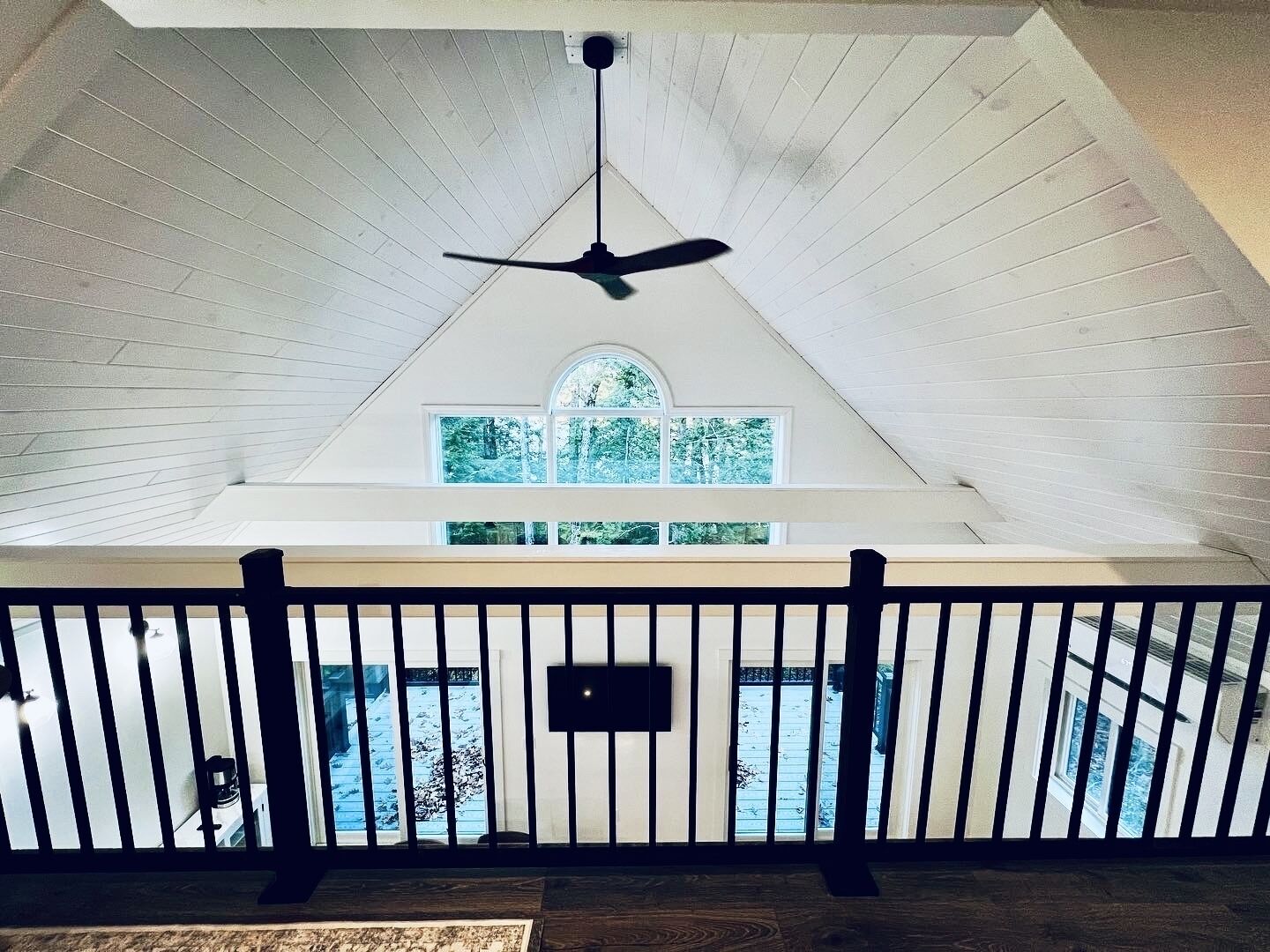 A room with a sloped ceiling, a ceiling fan, a window with a view of trees, and a railing.