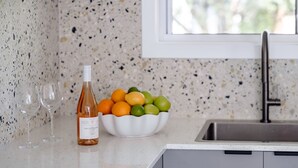 A kitchen counter with a bottle of wine, a bowl of fruit, and two wine glasses. - Little Miss Sunshine (Burrill Lake)