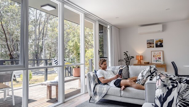 A modern living room with large windows, a wooden deck, and a sofa with decorative pillows. - Little Miss Sunshine (Burrill Lake)