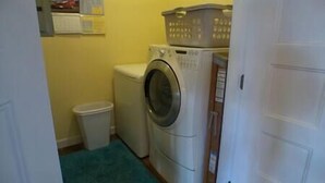 A compact laundry room with a washing machine, a small trash bin, and a blue rug. - Great Escape -2 Br 2Ba Waterfront Bay St Louis MS (Hancock County)