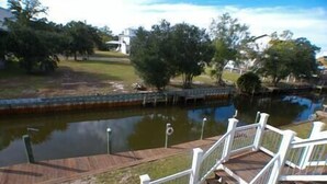 A wooden deck with white railings overlooking a calm body of water, surrounded by trees and a grassy area. - Great Escape -2 Br 2Ba Waterfront Bay St Louis MS (Hancock County)