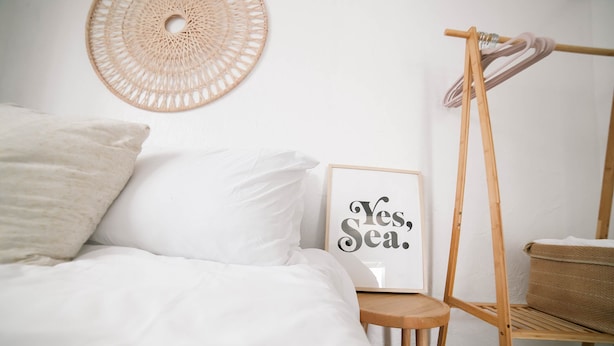 Un dormitorio ordenado con cama, almohadas, un cartel enmarcado, una silla de madera y una estantería con una cesta.