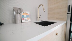 Una cocina con encimera blanca, un lavamanos negro, un grifo dorado y un juego de utensilios de cocina en un recipiente blanco.