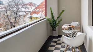 A balcony with a checkered floor, a potted plant, a small table with a chair, and a view of buildings with red roofs. - Luxury Piata Romana 2BR Urban Sanctuary by HOLT (Bucharest)