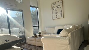 A modern living room with a white sofa, a small table with a plant, and a large window with a view of trees. - Luxe 2-Bedroom in Lavender Square (Langford)