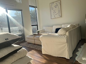 Ein modernes Wohnzimmer mit einem weißen Sofa, einem kleinen Tisch mit Pflanze und einem großen Fenster mit Blick auf Bäume.
