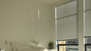 A bedroom with a bed, bedside tables, a nightstand, a window with blinds, and a view of the outside. - Luxe 2-Bedroom in Lavender Square (Langford)