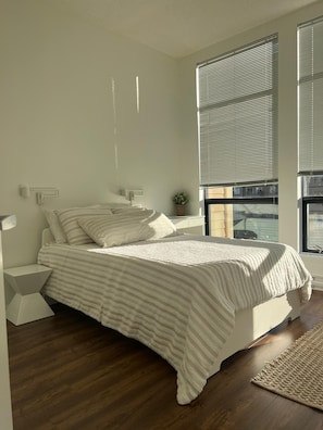 Ein Schlafzimmer mit Bett, Nachttischen, Fenster mit Jalousien und Blick ins Freie.
