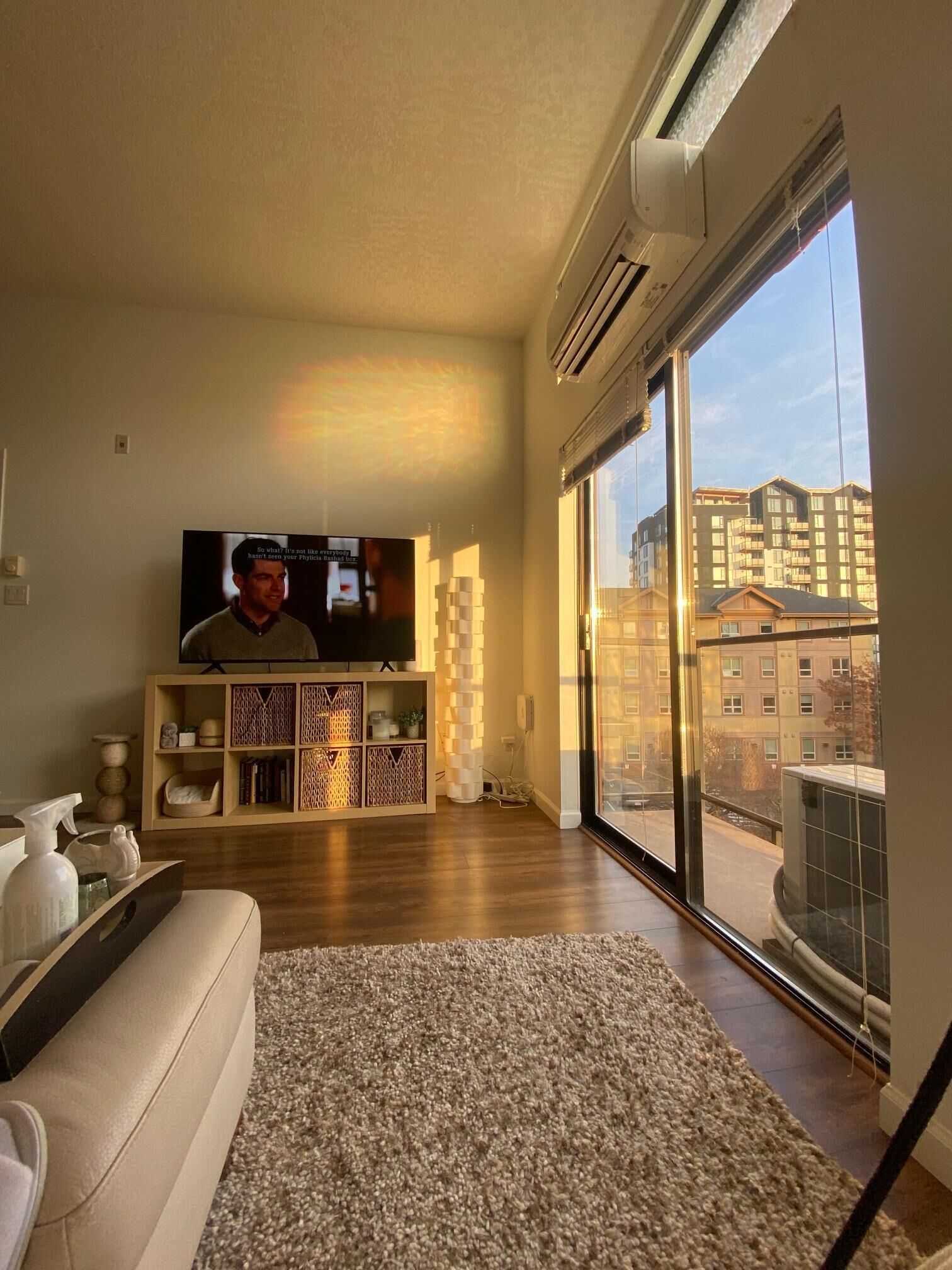 A living room with a large flat-screen TV, a wooden cabinet, a white sofa, and a large window offering a view of buildings.