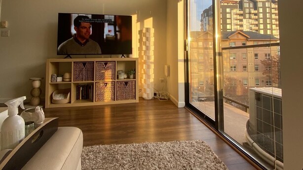 A living room with a large flat-screen TV, a wooden cabinet, a white sofa, and a large window offering a view of buildings. - Luxe 2-Bedroom in Lavender Square (Langford)