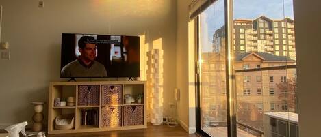 A living room with a large flat-screen TV, a wooden cabinet, a white sofa, and a large window offering a view of buildings.