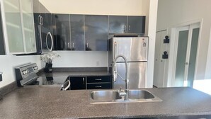 A kitchen with a stainless steel refrigerator, a double sink, and a built-in oven. There are three bar stools in front of the sink. - Luxe 2-Bedroom in Lavender Square (Langford)