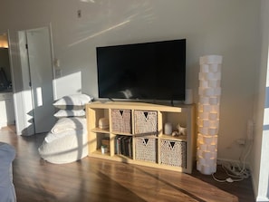 Ein Wohnzimmer mit einem Flachbildfernseher auf einem Holzboden, einer Reihe weißer Kissen und einer dekorativen Leuchte.