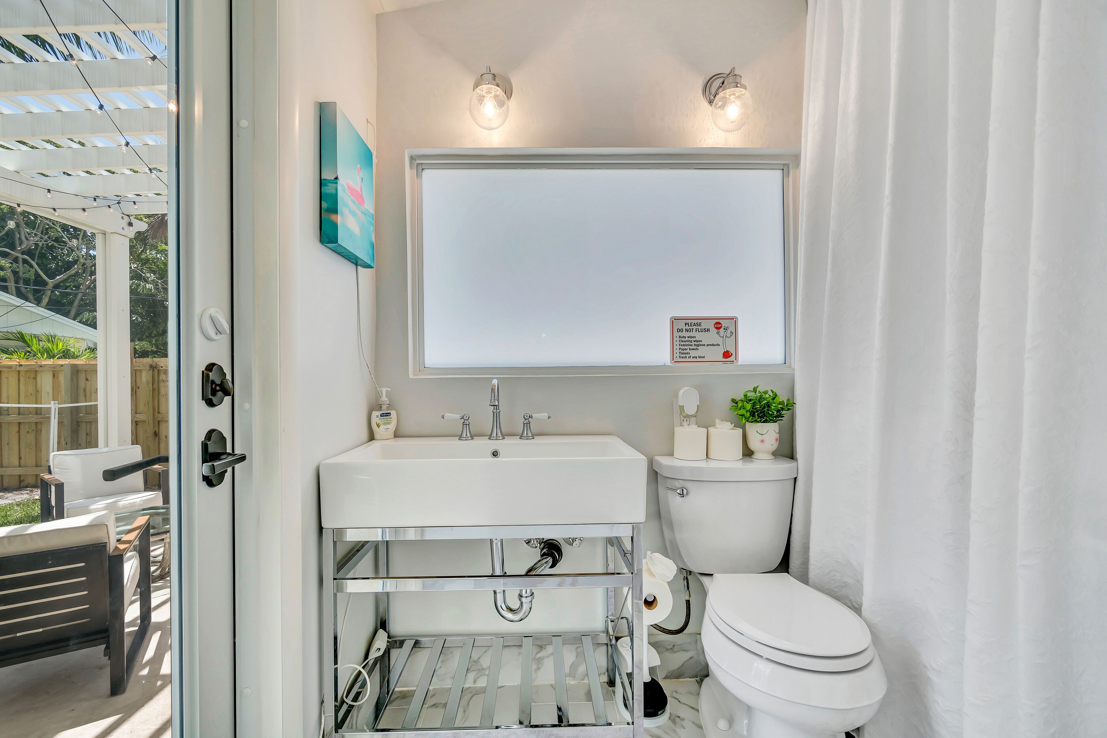 A compact bathroom with a white sink, toilet, and a large mirror above the sink. There is a small shelf with toiletries and a potted plant.