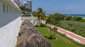 A thatched-roof structure adjacent to a white building, with a pathway leading to a beach and palm trees in the background. - Spacious Beachfront Retreat,
Family Friendly,
Free parking/Free Public Trolley (Miami Beach)