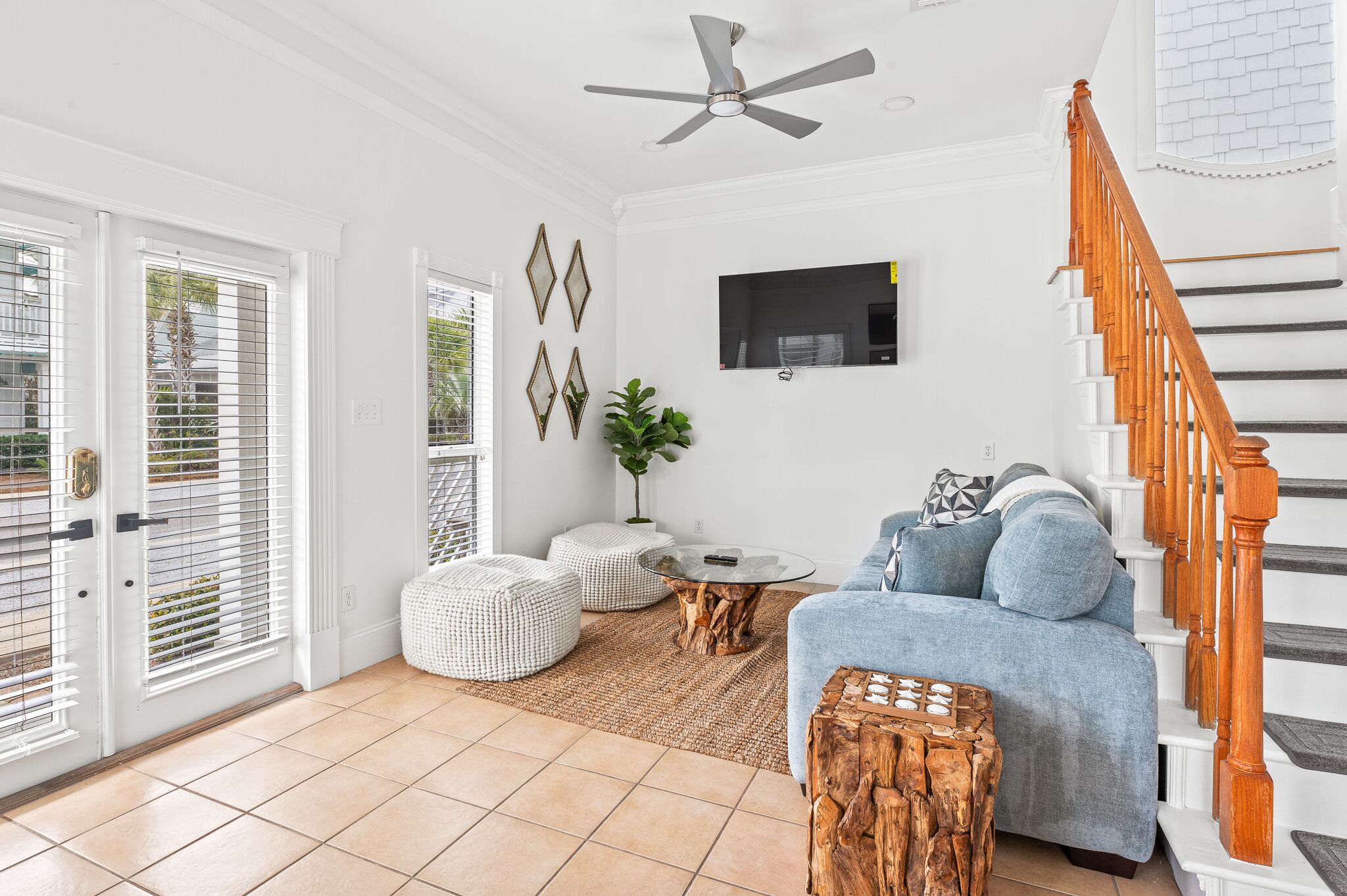 A spacious living room with a wooden staircase, a ceiling fan, a TV mounted on the wall, and a large window with blinds.