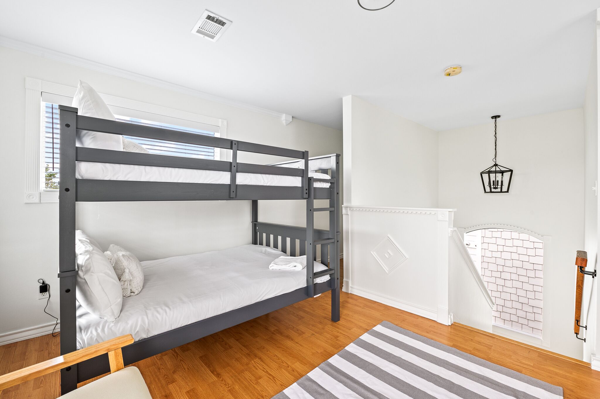 Une chambre avec un lit superposé, un plancher en bois, un mur blanc, une fenêtre et un luminaire suspendu.