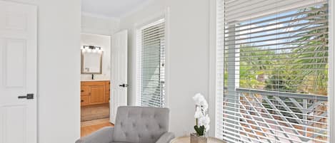 Een moderne woonkamer met een grijze bank, een kleine ronde tafel met een plant, en uitzicht door witte jaloezieën op een tuin.