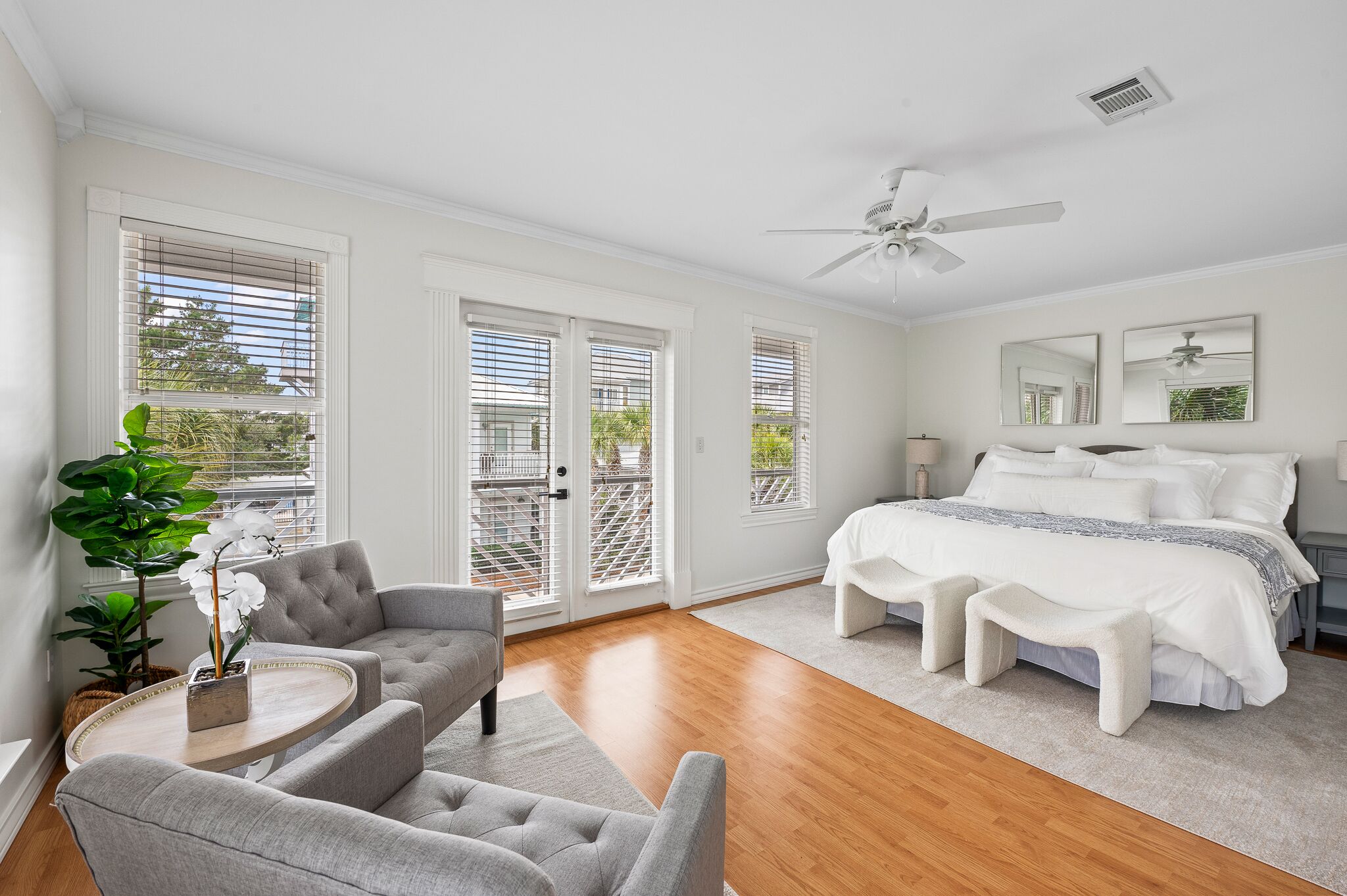 A bedroom with a bed, a bench, a grey sofa, a small table with a plant, and a ceiling fan.