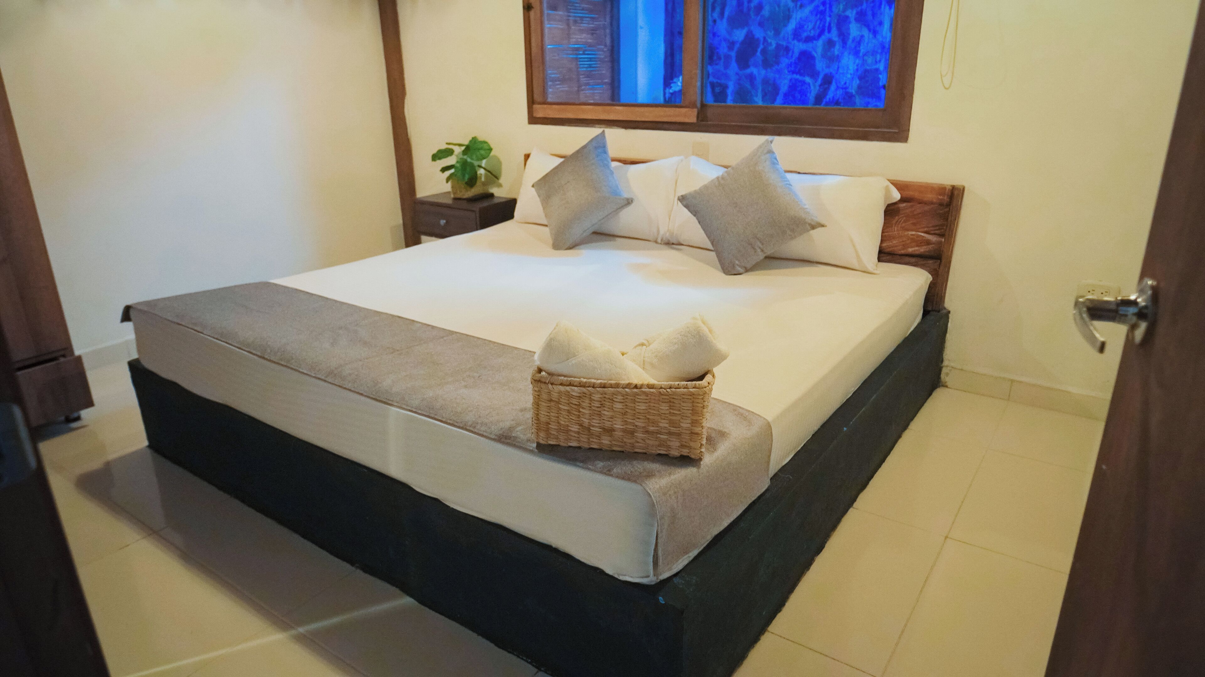A neatly made bed with a grey blanket and pillows, a wicker basket with towels, and a nightstand with a plant.