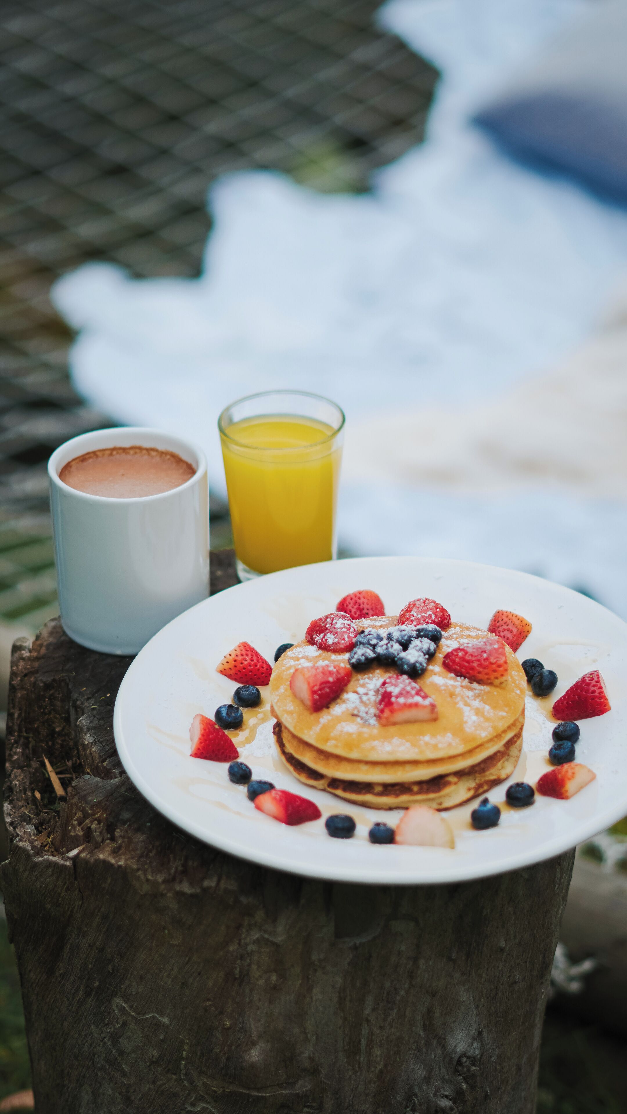 Ein Teller Pfannkuchen mit Erdbeeren und Blaubeeren, ein Glas Orangensaft und eine Tasse Kaffee auf einem hölzernen Baumstumpf.
