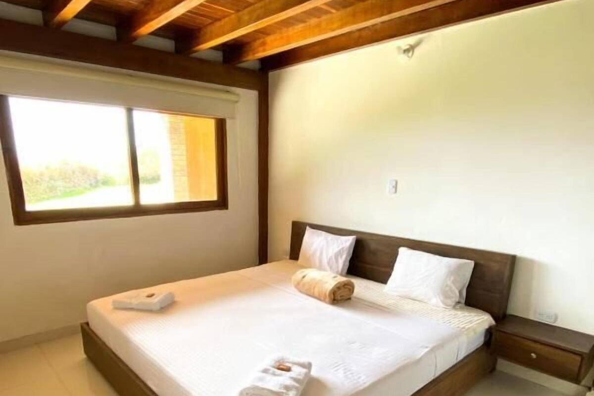 Una cama grande con cabecera de madera, una mesita de noche y una ventana con vistas a la vegetación.