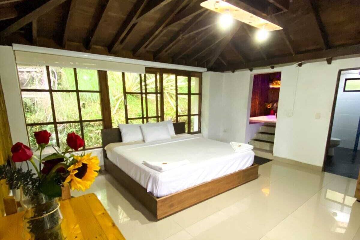 Un dormitorio con techo de madera, una cama grande, una mesa de madera con un florero y vistas a zonas verdes a través de las ventanas.