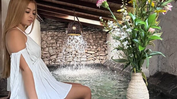A woman in a white dress sitting by a stone wall with a water feature and a vase of flowers.