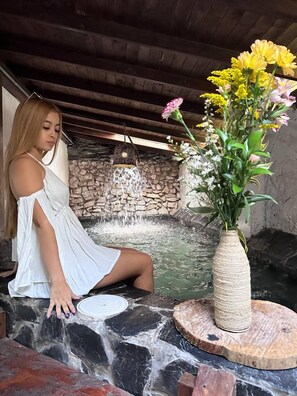 Eine Frau in einem weißen Kleid sitzt an einer Steinmauer mit einem Wasserspiel und einem Blumenstrauß.