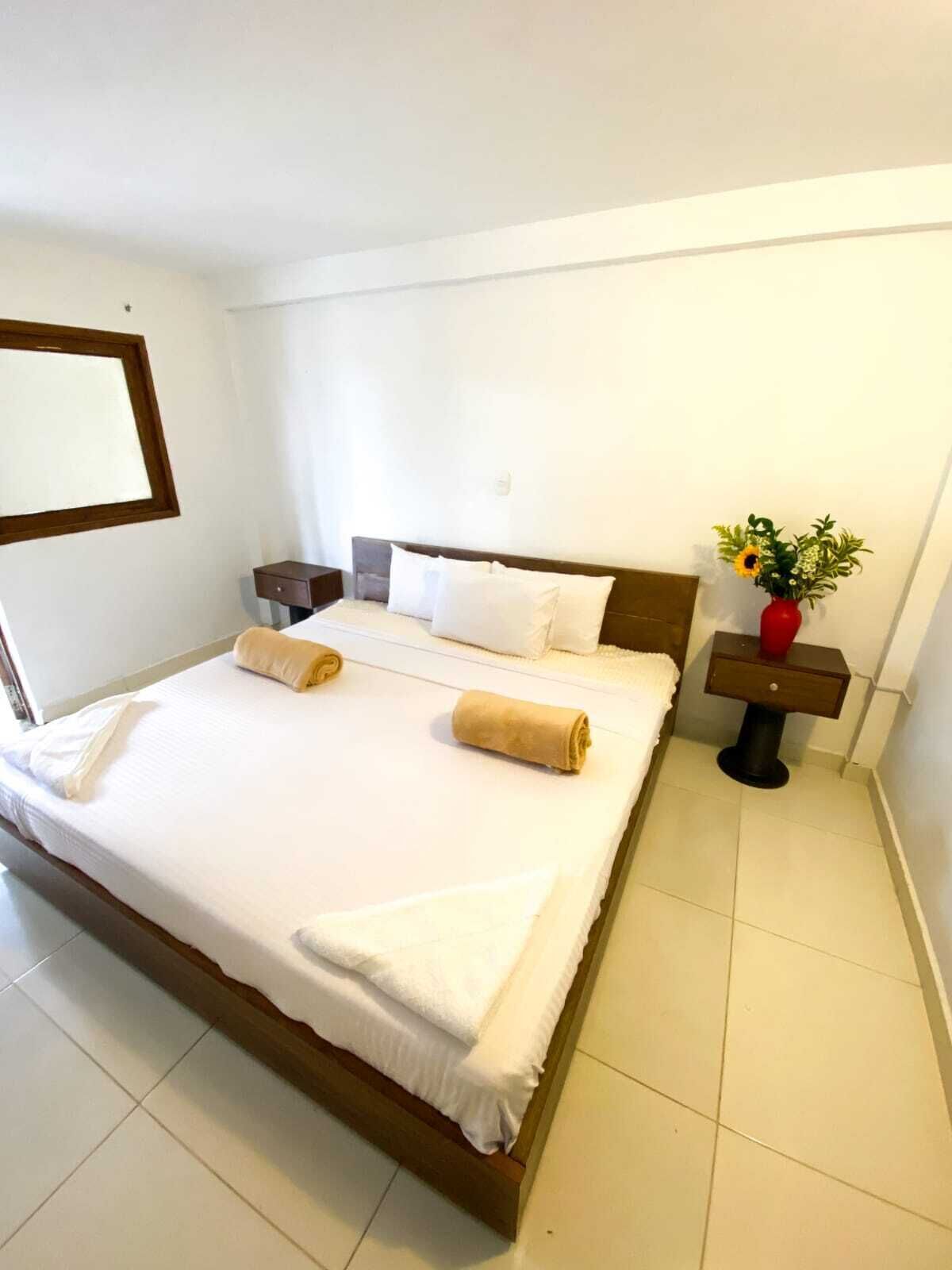 Una cama bien hecha con sábanas blancas y dos toallas enrolladas, una mesita de noche con un florero y un espejo montado en la pared.