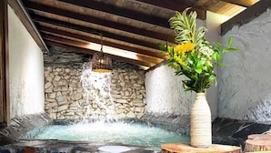 A stone hot tub with a waterfall feature, wooden steps, and a vase with flowers.