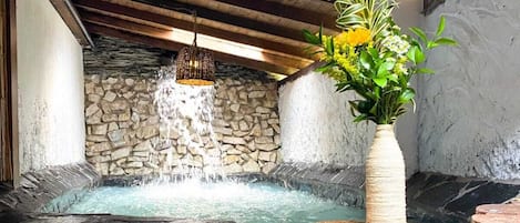 A stone hot tub with a waterfall feature, wooden steps, and a vase with flowers.