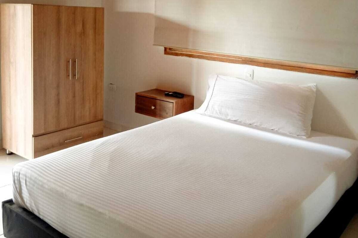 Un dormitorio con techo de madera, una cama con ropa de cama blanca, un armario de madera y una mesita de noche con control remoto.