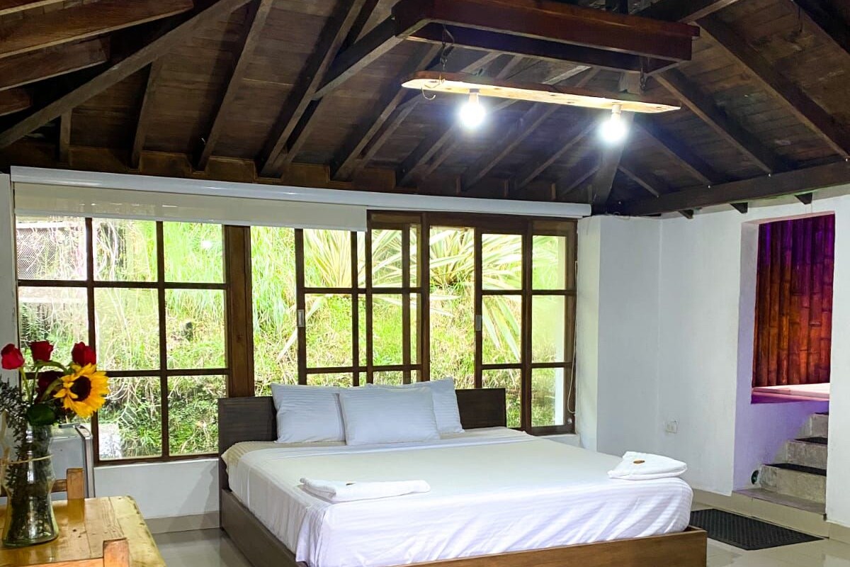 Une chambre à coucher avec un grand lit, une table en bois ornée d ’un vase de fleurs et une vue sur la verdure grâce à de grandes fenêtres.