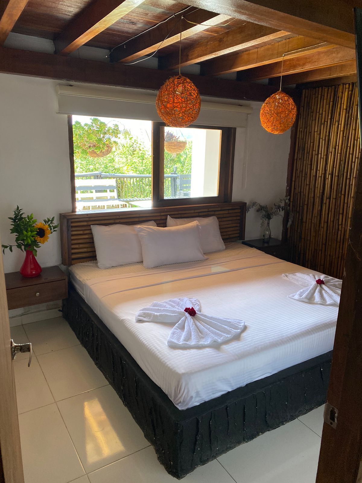 A hotel room with a large bed, wooden headboard, two hanging orange lamps, a window with a view of greenery, and a red vase with flowers on a nightstand.