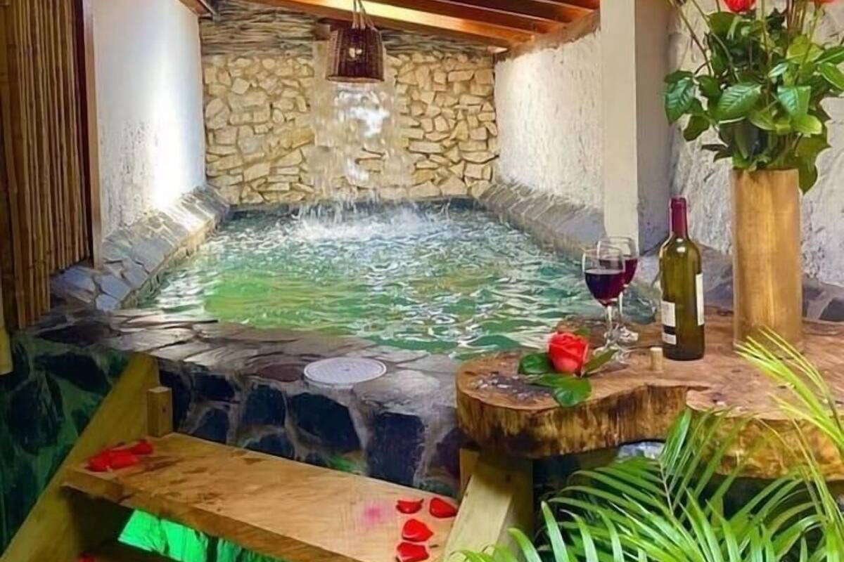 Un jacuzzi con paredes de piedra y vigas de madera, rodeado de follaje y un jarrón de flores.