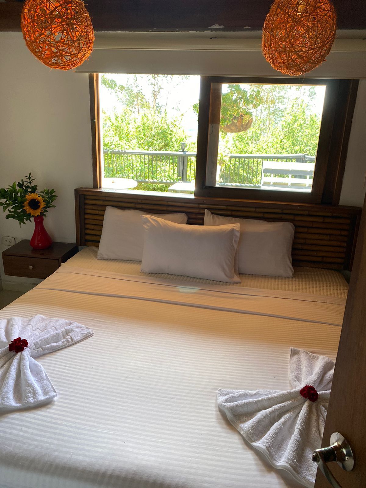 A hotel room with a bed, two pillows, a nightstand with a red vase and a sunflower, and two hanging lamps.