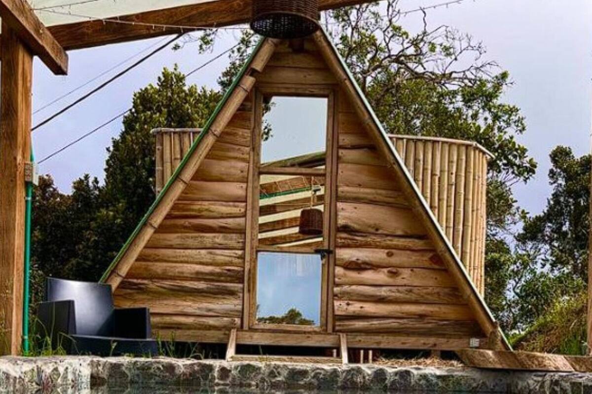 Una cabaña de madera con techo triangular, un balcón pequeño y una piscina cristalina al frente.
