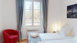 A hotel room with a bed, a chair, a window with curtains, a radiator, and a wall-mounted picture.