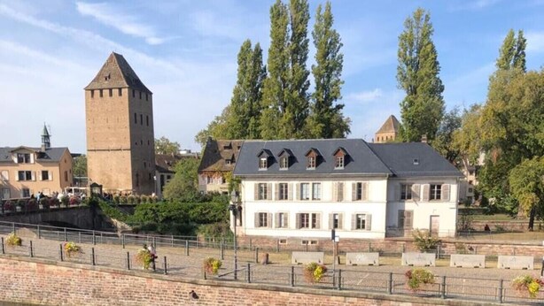 A riverside view of a historic building with a tower, a large white house with multiple windows, and a stone wall.
