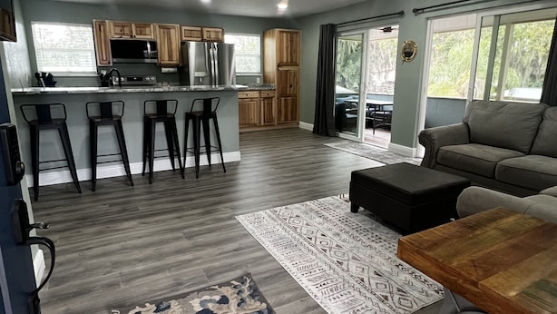 A modern kitchen with wooden cabinets, a central island with bar stools, a dining area with a sofa and ottoman, and a ceiling fan.